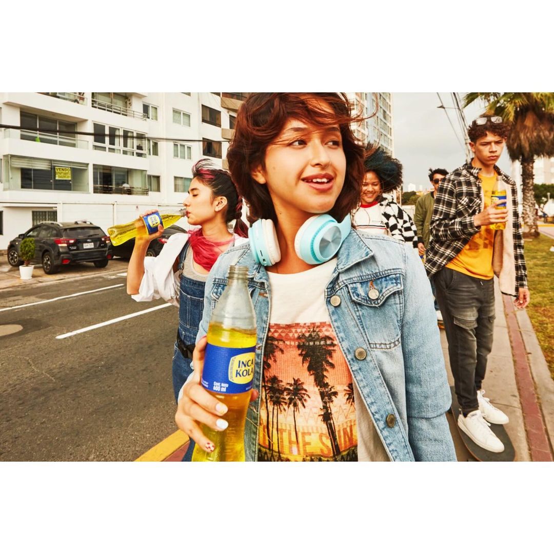 Several young adults walk down a city street holding bottles of Inca Kola soda, wearing casual summer outfits and headphones.