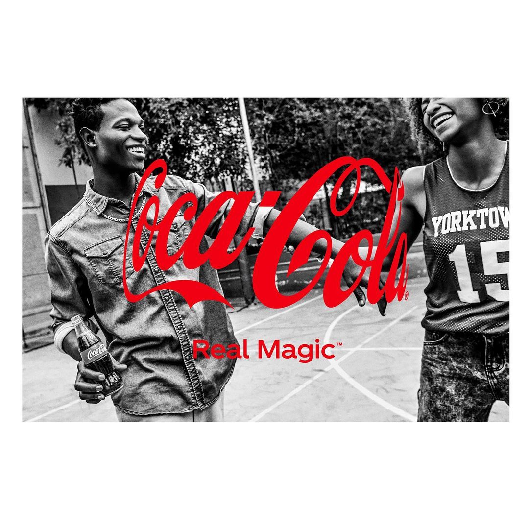 Two smiling young adults in black and white hold a Coca-Cola bottle, with the Coca-Cola logo and Real Magic tagline in red.
