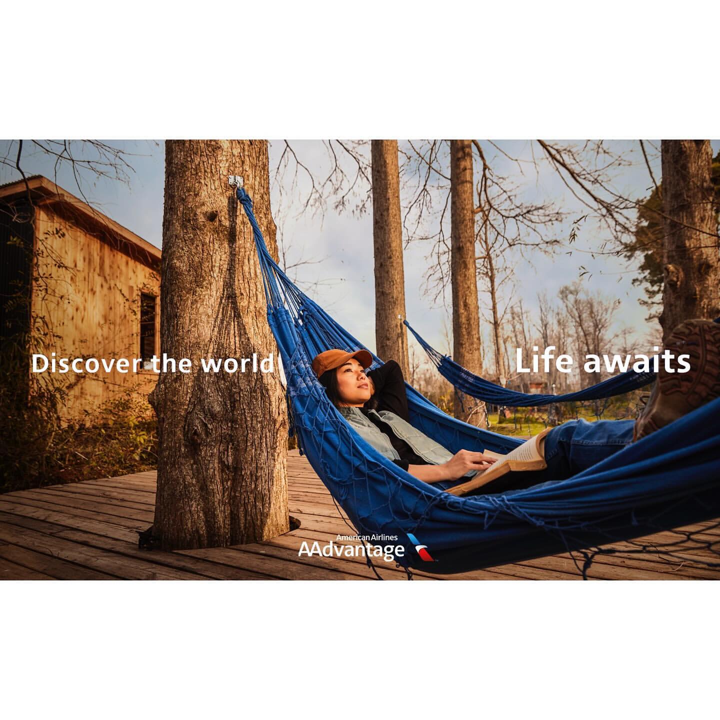 Woman relaxing in a blue hammock strung between trees, with a glimpse of a rustic building and an airline logo visible.