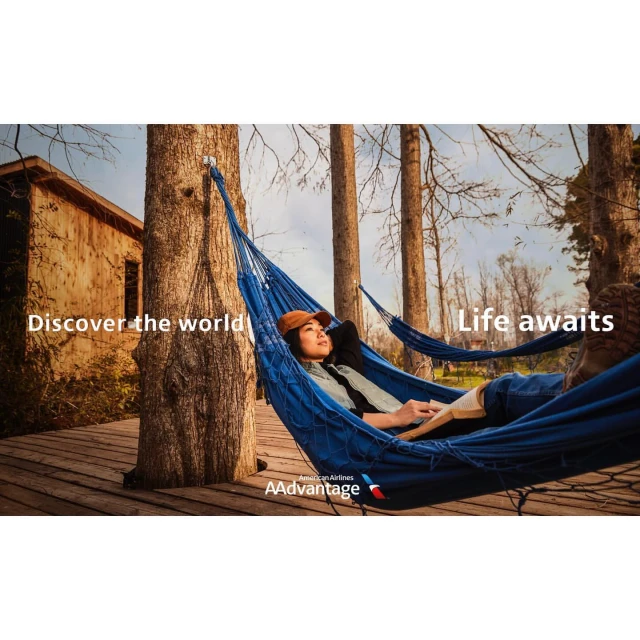 Woman relaxing in a blue hammock strung between trees, with a glimpse of a rustic building and an airline logo visible.