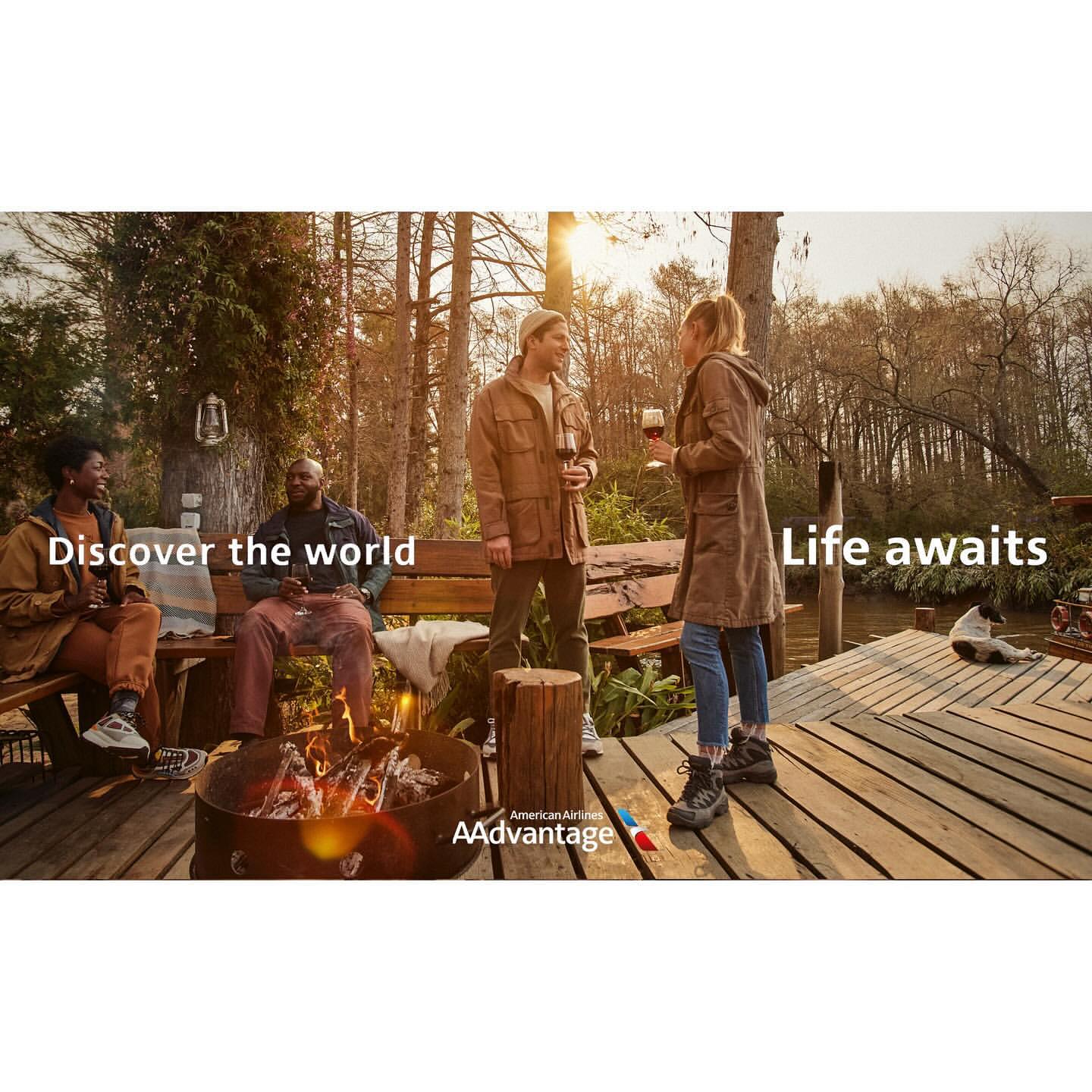 Group of diverse friends sitting around a fire pit near a lake, holding wine glasses while trees and sunlight fill the background.