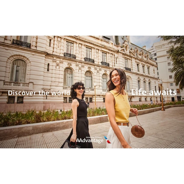 Two adult women smile, walking along a sidewalk in front of a European-style building on a sunny day with blue skies.