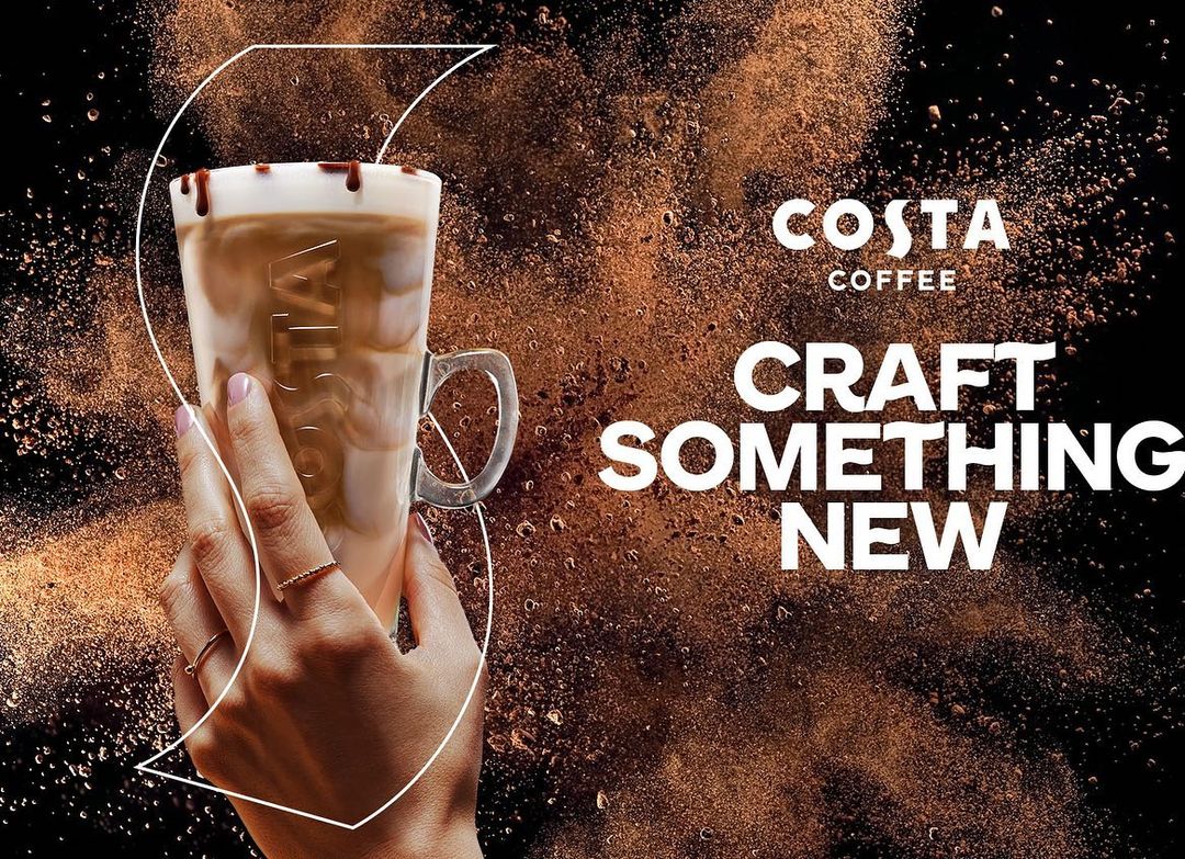 Costa Coffee ad: Hand holds a glass of coffee with chocolate drizzle on a dark background with coffee bean dust and text.