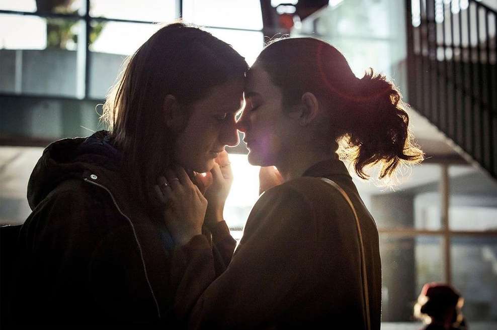 Intimate portrait of two young women with foreheads touching in a sun-drenched urban indoor setting, captured in soft focus.