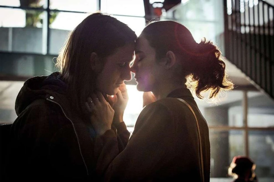 Intimate portrait of two young women with foreheads touching in a sun-drenched urban indoor setting, captured in soft focus.