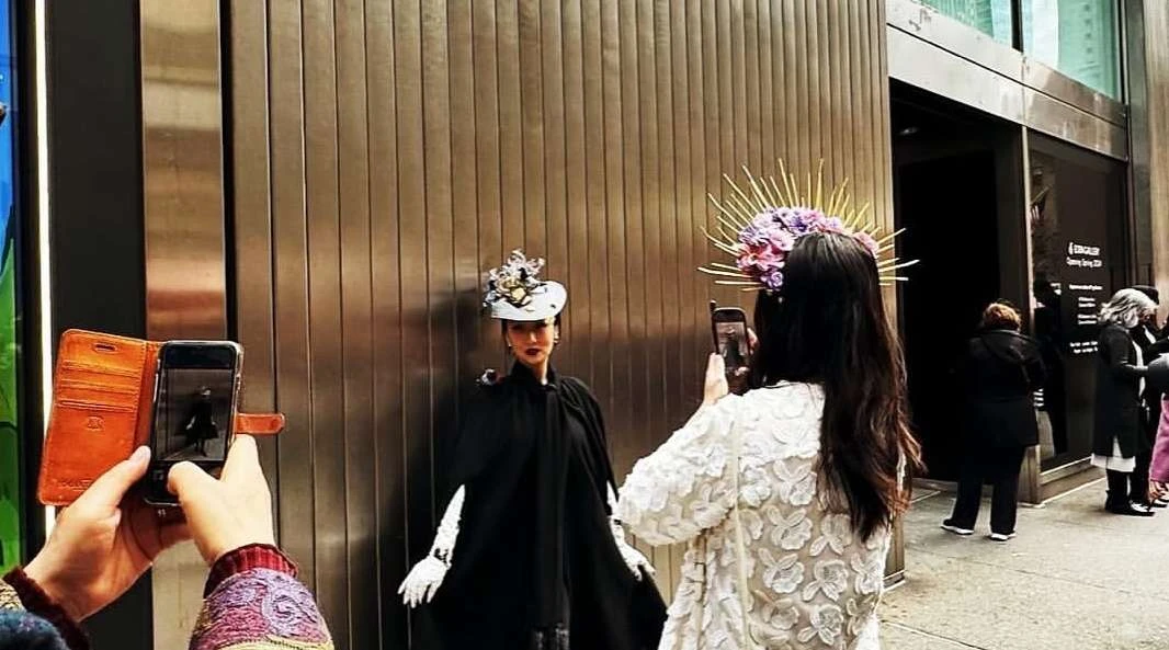Two women in formal attire and hats pose against a metal wall as other people take pictures with their phones outdoors.