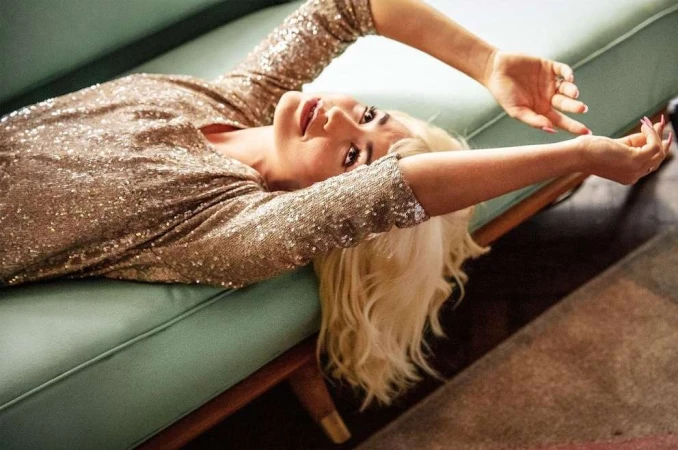 Blonde woman wearing a sparkling gold sequin dress lying back on a mint green sofa.