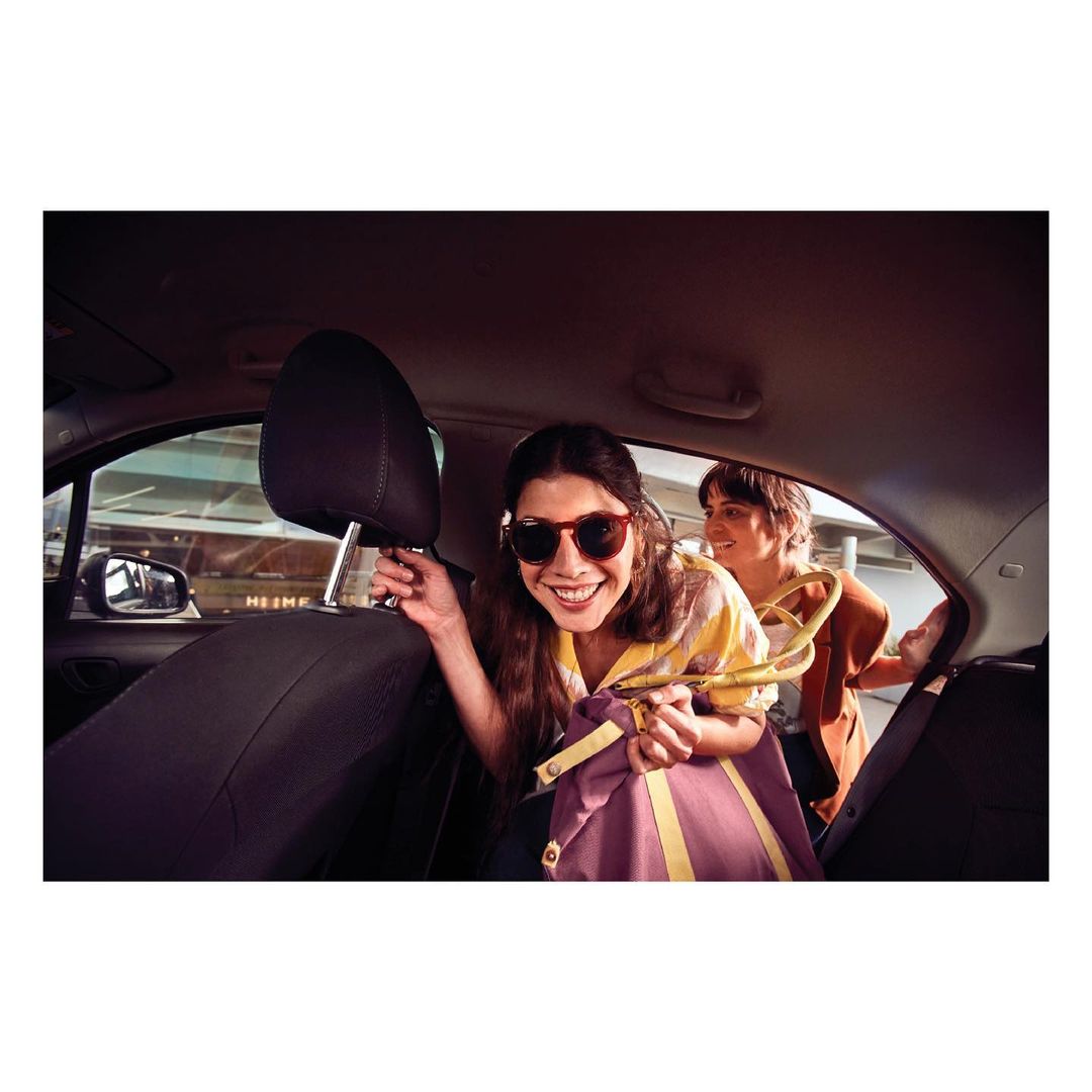 Two adult women are sitting in the back of a car; one smiling with sunglasses and holding a shopping bag, hanger with clothing in the background.