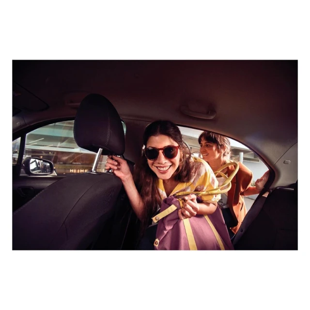 Two adult women are sitting in the back of a car; one smiling with sunglasses and holding a shopping bag, hanger with clothing in the background.