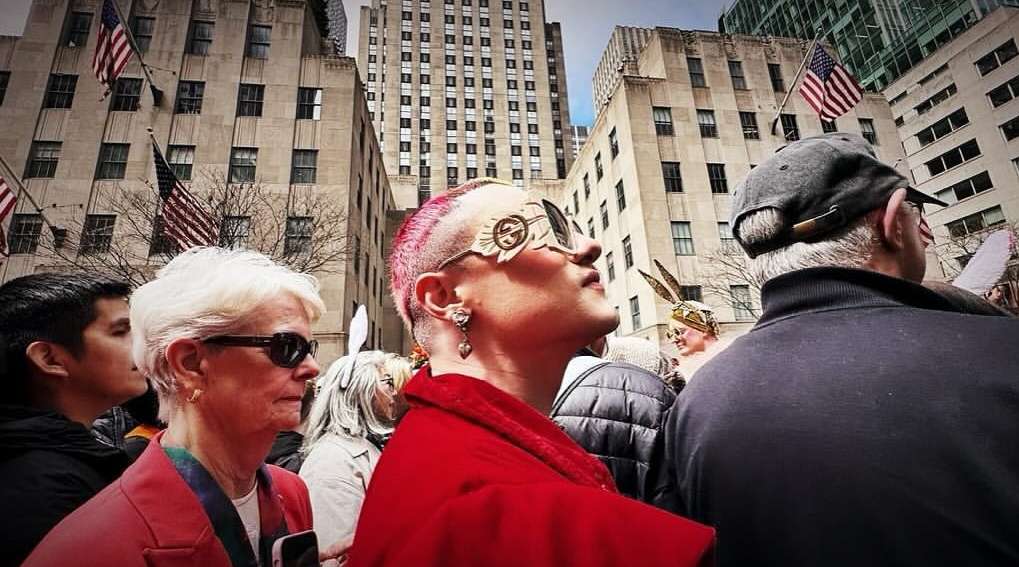 Eye-level outdoor shot of a diverse crowd in front of urban buildings with American flags; a woman with pink hair is in focus.