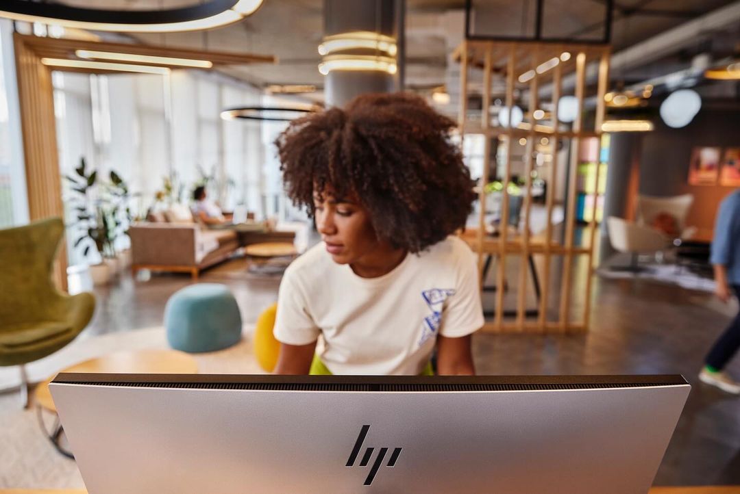 Woman with afro working on a computer in an office lounge area with chairs, tables, and architectural details visible in soft light.