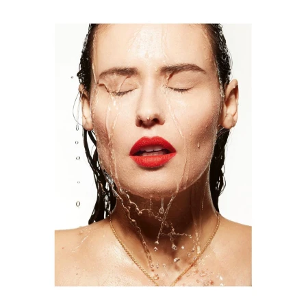 Close-up of a woman's face with closed eyes and red lips, water cascading down her face against a bright white backdrop in a studio setting.