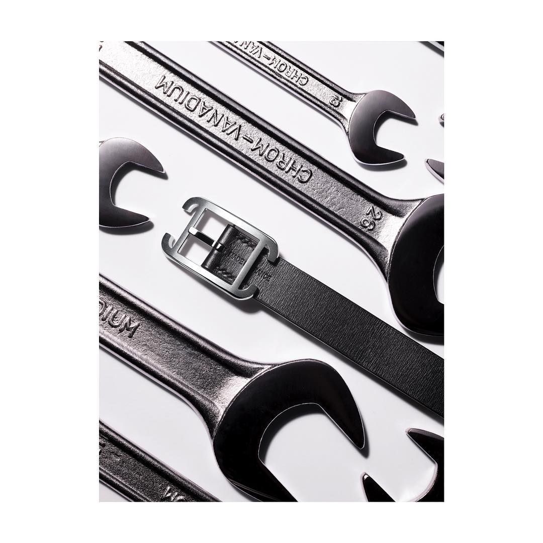 Close-up of a black leather belt with a silver buckle placed diagonally over an arrangement of silver chrome vanadium wrenches.