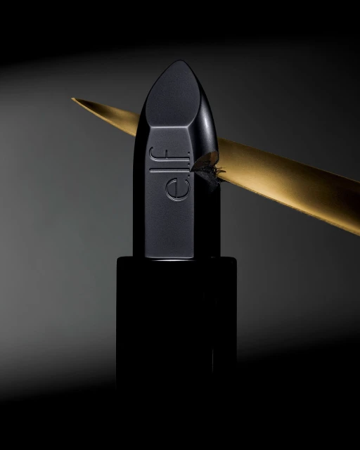 A black lipstick from e.l.f. Cosmetics is presented with a gold accent on a smooth grey gradient background in a product photography style.