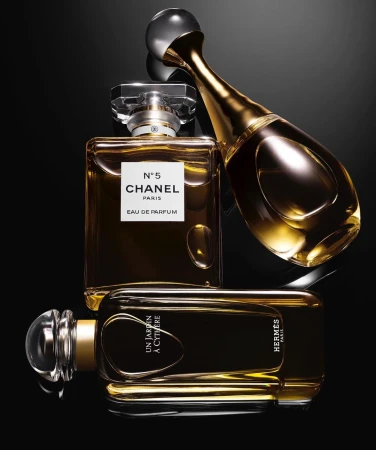 Three perfume bottles sit on a black reflective surface. The bottles are Chanel No. 5, Hermes Un Jardin A Cythere, and Dior J'Adore.