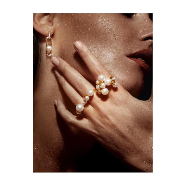 Close-up of woman's face and hand wearing pearl and gold jewelry, including a ring and earrings, with dewy skin.