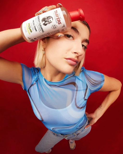 Young woman poses with a Thayers facial toner bottle in a studio, set against a vibrant red backdrop, showcasing beauty and skincare product.