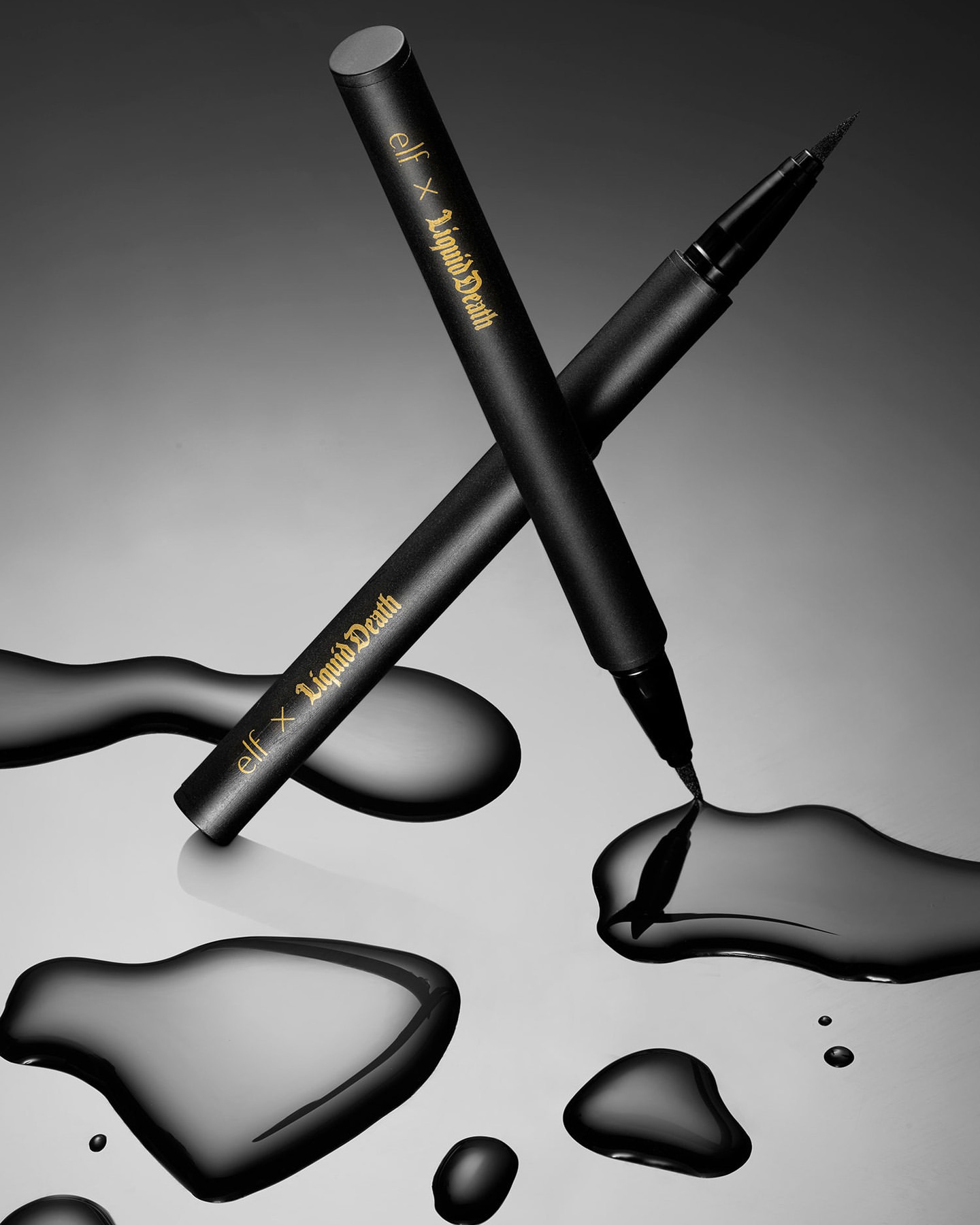 Two black e.l.f. x Liquid Death eyeliner pens, one with the cap on, rest on a reflective surface with drops of liquid eyeliner.