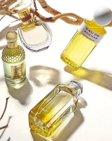 Four perfume bottles from Rochas, Hermes, and Guerlain brands arranged on a white surface with light tan-colored sticks.