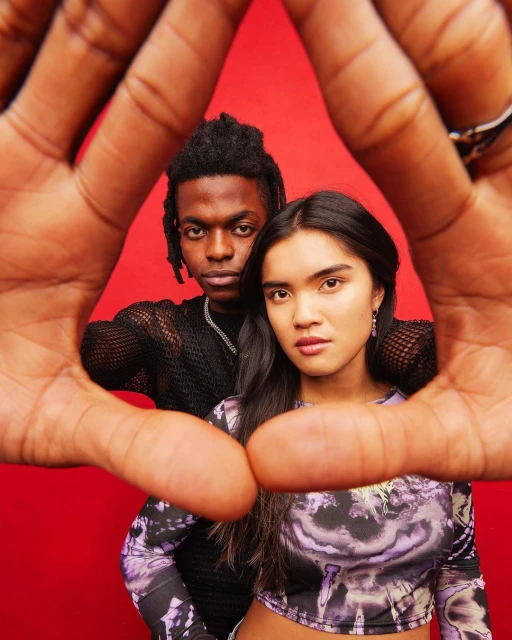 Close-up of an African-descent man and Asian-descent woman posing against a red background, framed by a human-made triangle with hands.