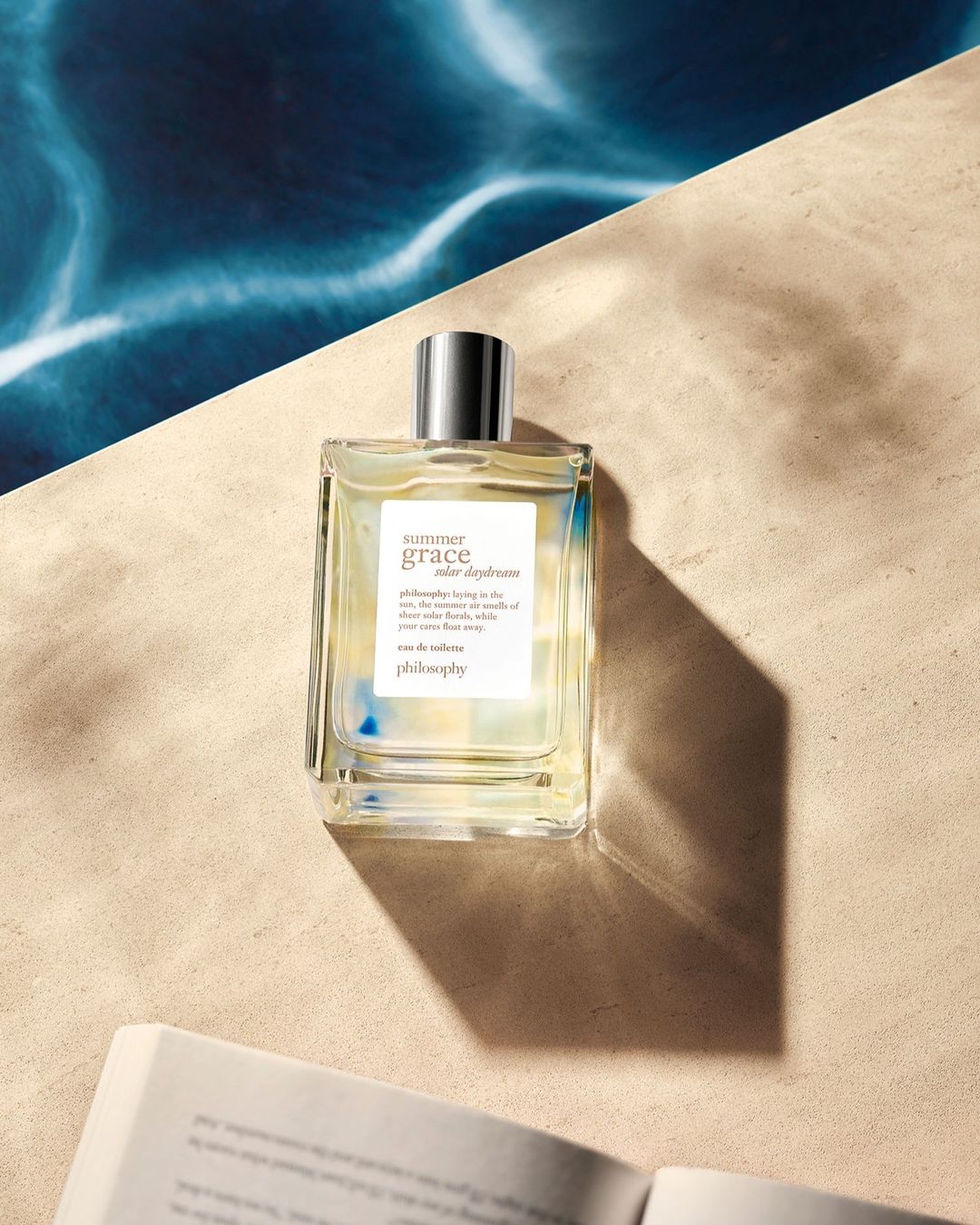 Still life of a glass perfume bottle with a silver cap on tan surface, set against blue water design and open book.