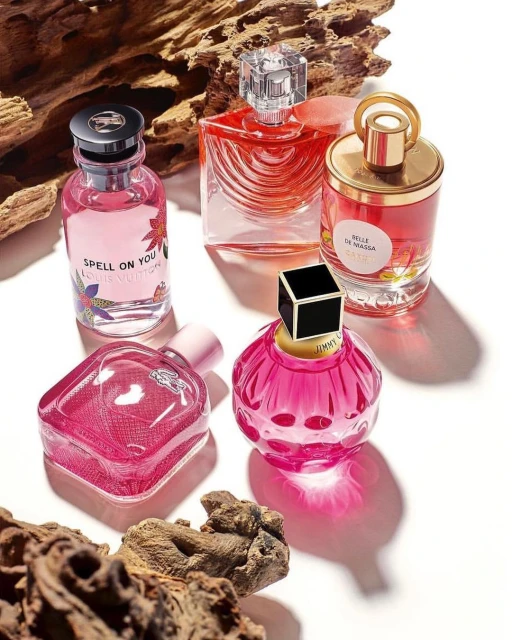 Five perfume bottles of various shapes and sizes are arranged on a light surface with textured wood pieces scattered around them.