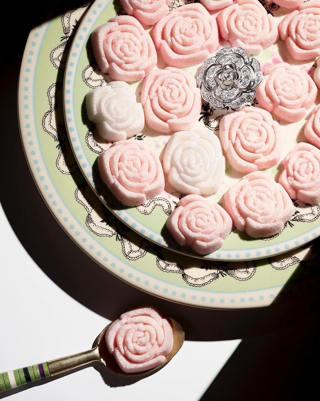 Overhead studio shot of rose-shaped sugar candy in pastel pink and white shades, arranged on decorative plates, plus a silver flower ring.