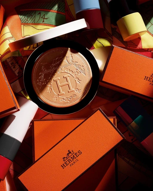 Hermès makeup compact in black case with embossed logo. Cosmetic surrounded by orange Hermès boxes and nail polish.