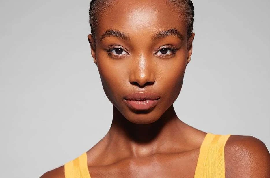 Close-up portrait of a black female adult with dark eyes and shiny lips, wearing a yellow tank top against a grey background.