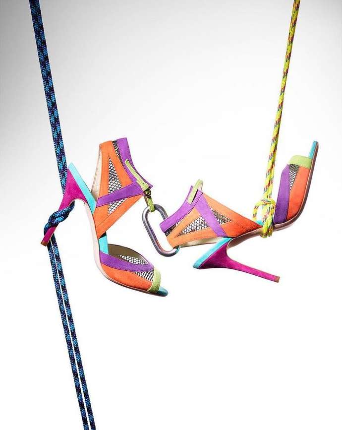 Vibrant heeled shoes suspended by ropes and a carabiner against a white background, showcasing fashion and climbing gear.