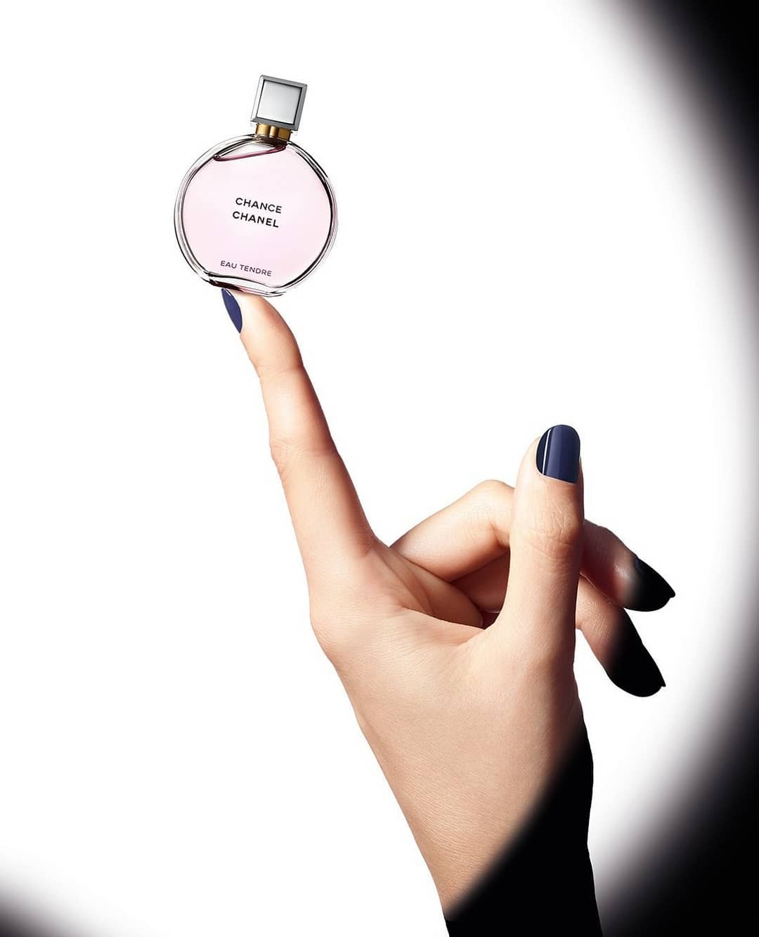 Close-up studio shot of a perfume bottle balanced on a woman's fingertip, set against a white background with dark, diffused shadows.