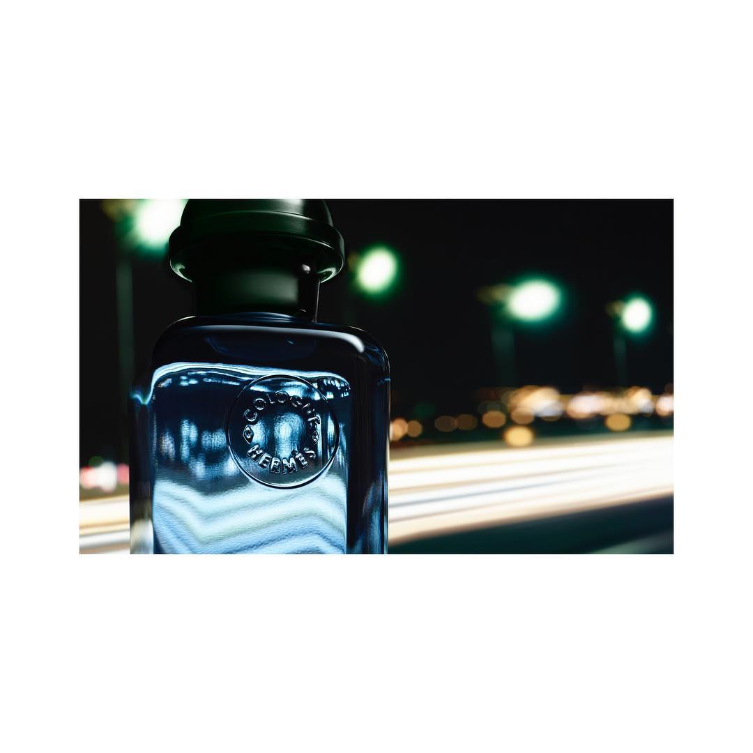 Cologne bottle by Hermes stands in front of blurred city lights at night, dark blue and black gradients, studio product shot.