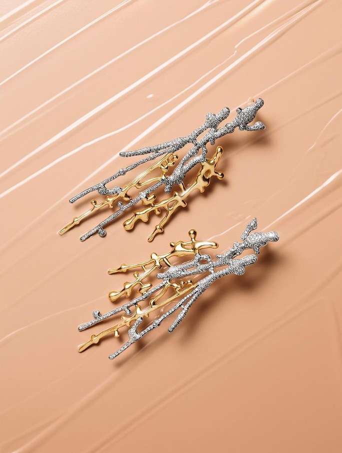 A pair of abstract earrings made of silver and gold metal are displayed on a beige surface, illuminated with soft lighting for a high-fashion look.