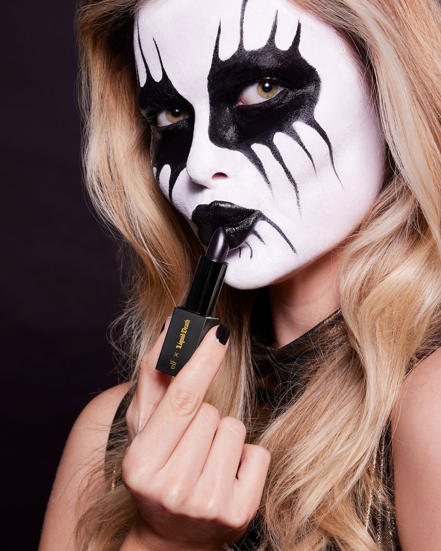 Woman in black and white face makeup holding a black lipstick tube in a studio with a dark background for a beauty shot.