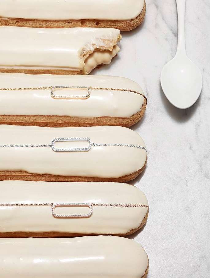 Close-up of white-frosted eclairs decorated with gold, silver, and rose-gold chain necklaces, arranged on a marble surface with a white spoon.