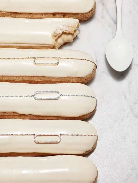Close-up of white-frosted eclairs decorated with gold, silver, and rose-gold chain necklaces, arranged on a marble surface with a white spoon.