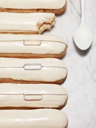 Close-up of white-frosted eclairs decorated with gold, silver, and rose-gold chain necklaces, arranged on a marble surface with a white spoon.