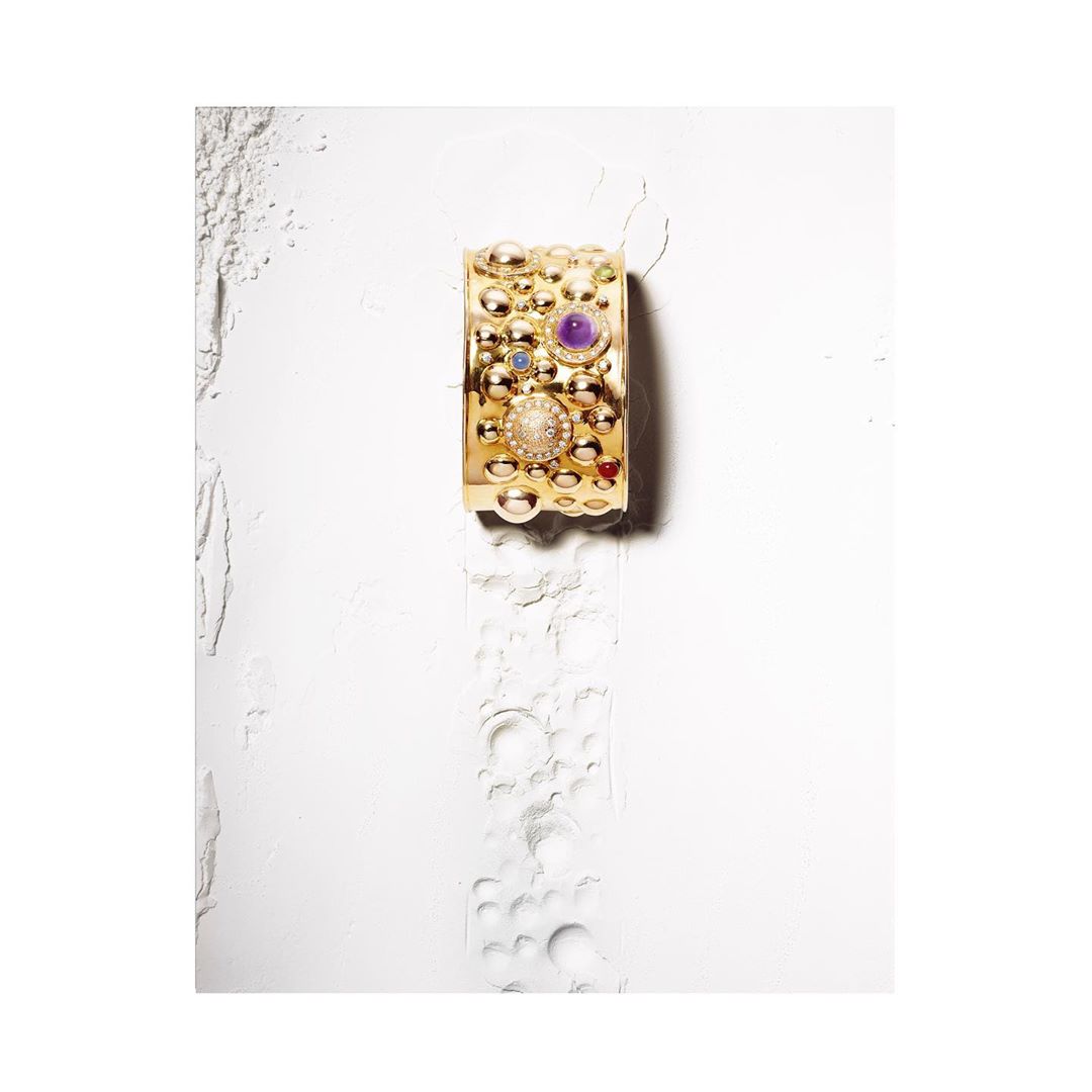 Close-up still life of an ornate gold cuff bracelet with gemstones on a textured white surface. Studio product shot.