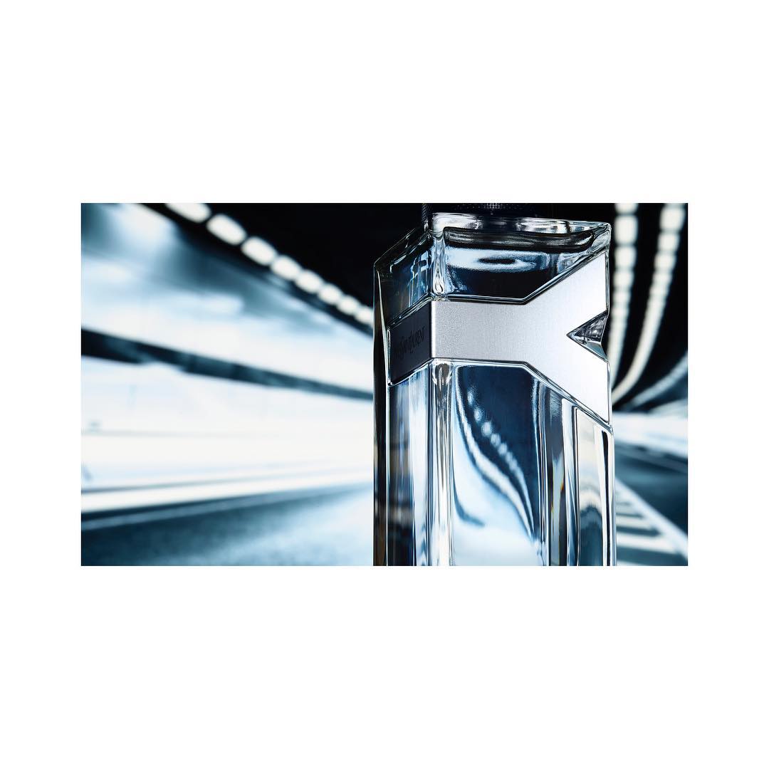 Close-up of a modern glass fragrance bottle with a silver design against a blurred background with light trails. Product shot for advertising.