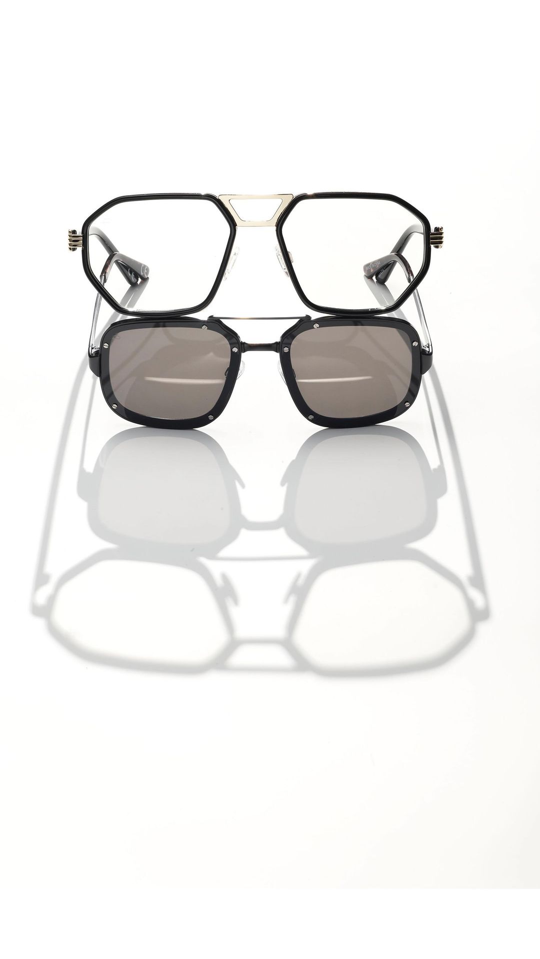 Studio shot of two pairs of stylish eyeglasses stacked, one with clear lenses and the other with sunglasses, casting strong shadows.