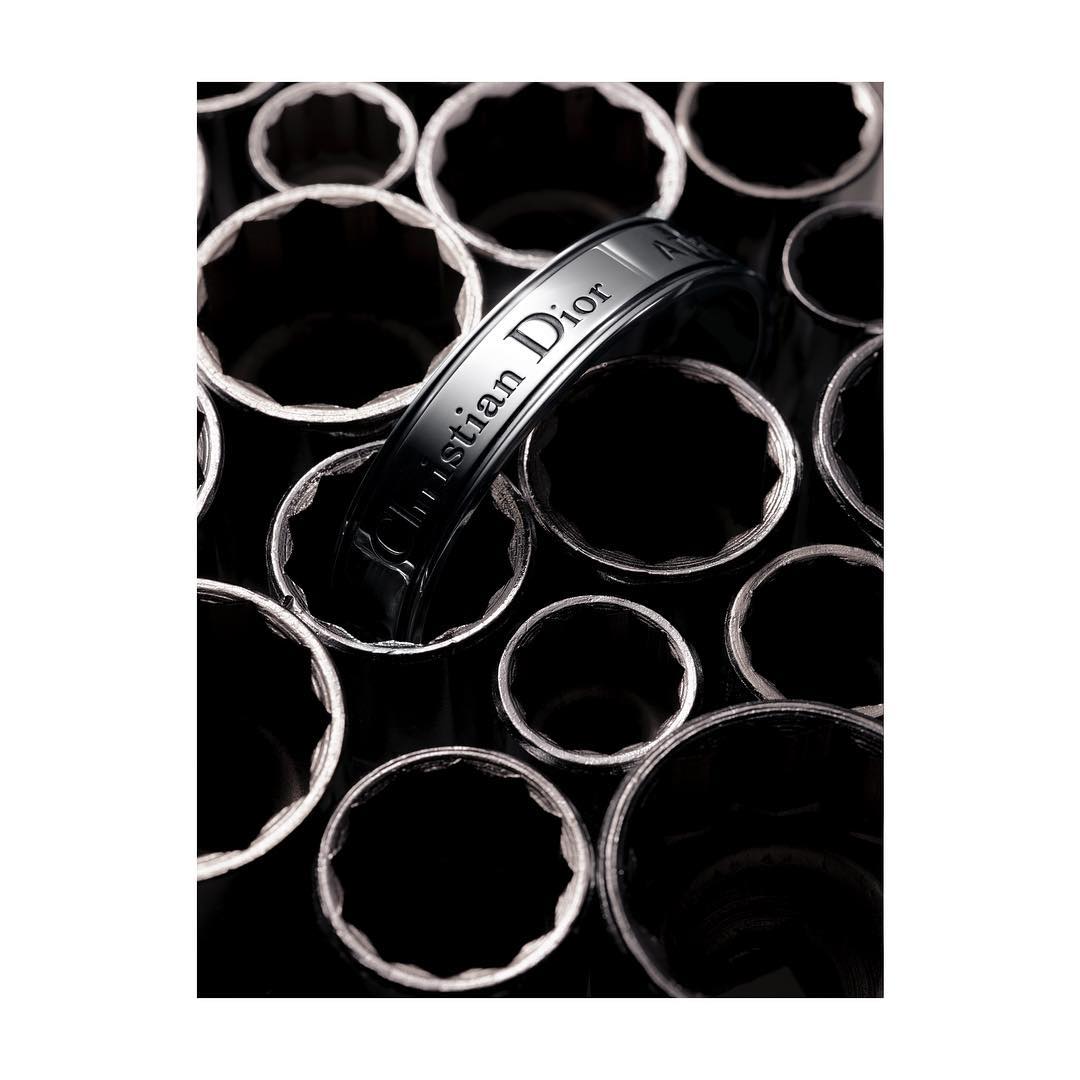 Close-up of a black 'Christian Dior' branded ring on a black reflective surface, surrounded by metallic, toothed rings.