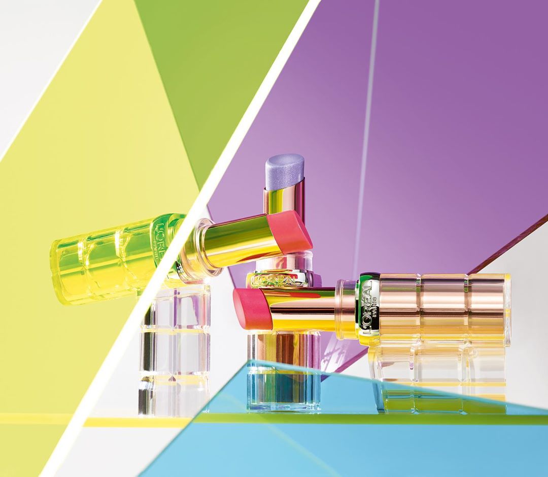 Still life of L'Oreal lipsticks with iridescent gold tubes and clear bases, arranged on a surface with geometric color blocks.