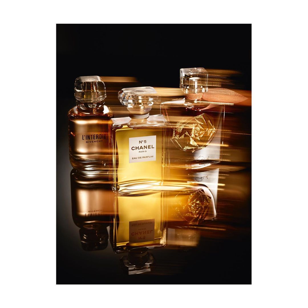 Three perfume bottles, including Chanel N°5, glisten on a reflective black surface against a blurred golden and black background. 