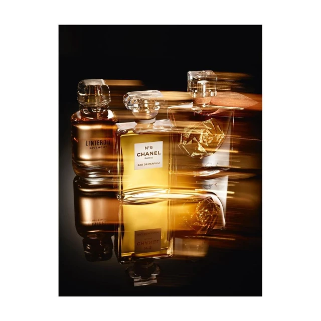 Three perfume bottles, including Chanel N°5, glisten on a reflective black surface against a blurred golden and black background. 