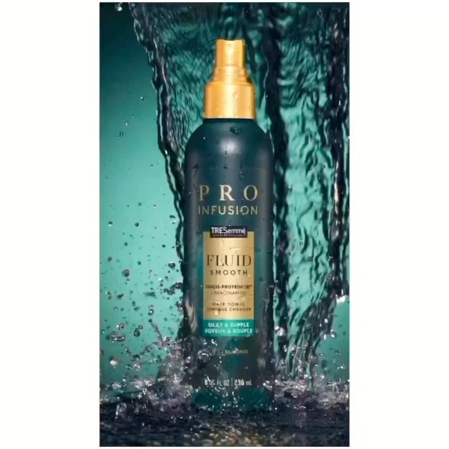 Dark teal Tresemme Pro Infusion Fluid Smooth hair tonic spray bottle with gold pump, against an out-of-focus teal water background.