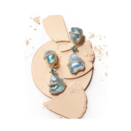 Close-up product shot of iridescent baroque pearl earrings resting on shattered beige cosmetic powder fragments against a white background.