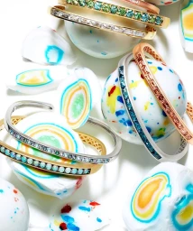 Close-up product photography of gemstone and gold bangles resting on colourful candy-like spheres against a bright white background.