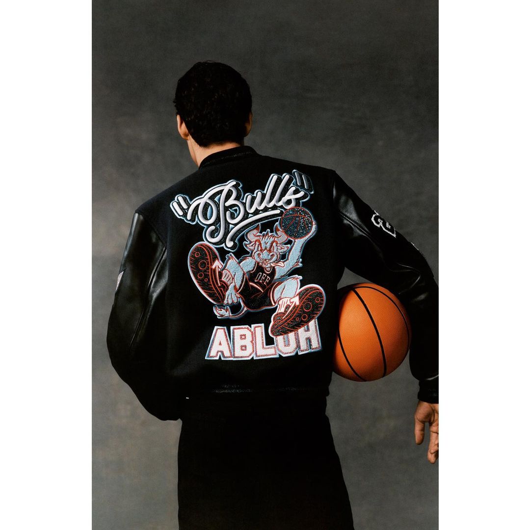 Rear view of an adult male wearing a black jacket with a cartoon graphic and the word Bulls printed on the back, holding a basketball.