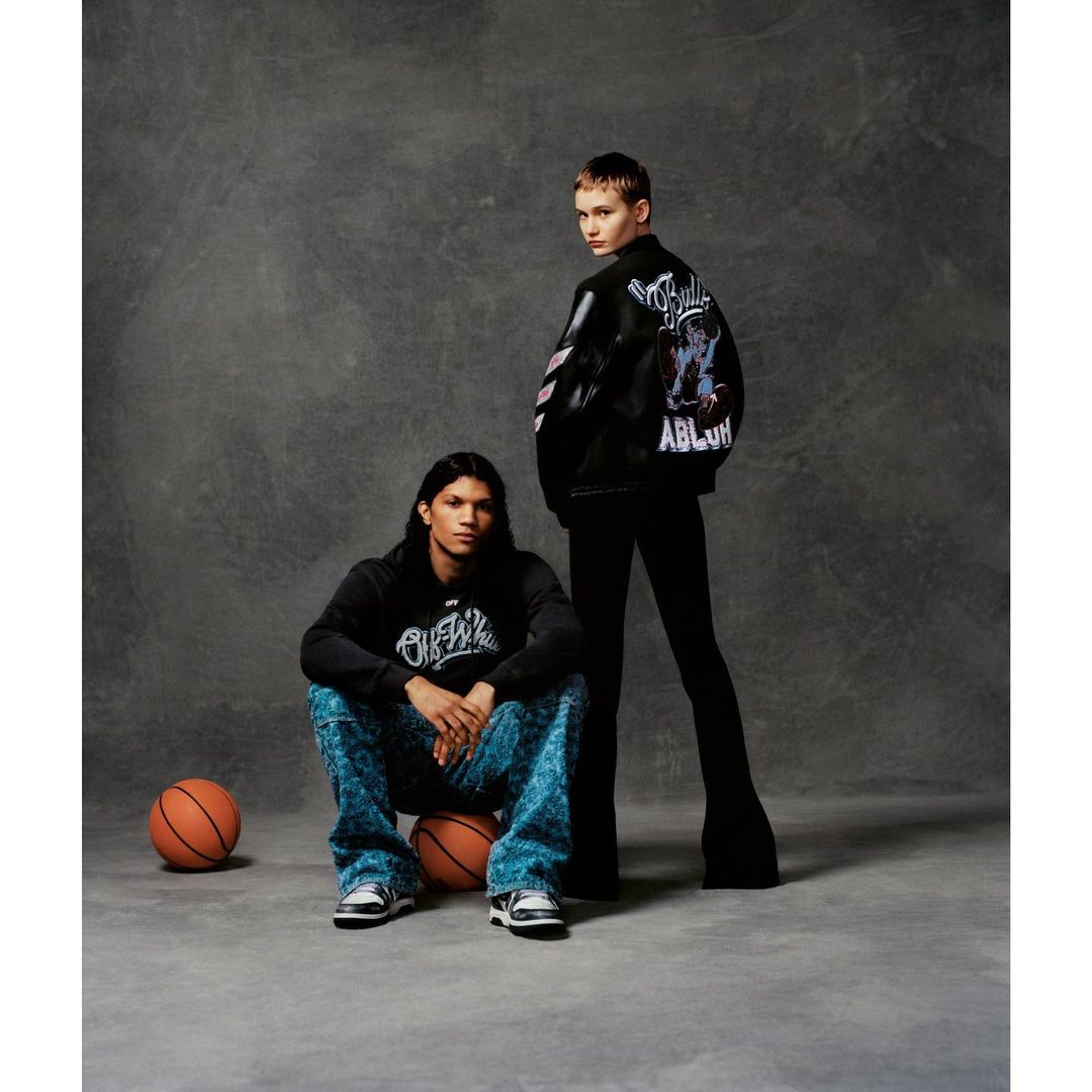 Two fashion models pose in a studio setting with basketballs. One model sits on the basketballs, and the other stands behind them.