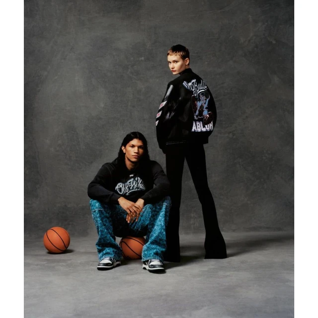 Two fashion models pose in a studio setting with basketballs. One model sits on the basketballs, and the other stands behind them.